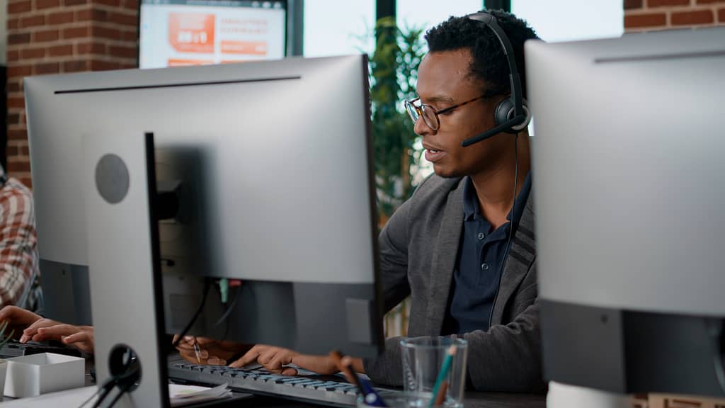 African american employee using headset at customer service job, answering call about telemarketing sales. Male operator working at call center office to help clients on helpline.